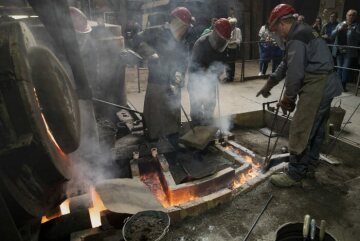 Arbeiter mit Helm und Gesichtsschutz arbeiten an Gießkanälen neben dem glühenden Schmelzofen bei der Produktion einer Glocke, am 22. April 2022 in der Werkstatt der Glocken- und Kunstgießerei Rincker in Sinn.