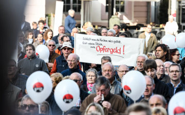 Demonstranten mit einem Plakat mit der Aufschrift "Schließt man die Kirchen wies gefällt, fehlt unterm Strich, auch Opfergeld!" bei der "Protestaktion "5 vor 12" gegen Großpfarreien der Initiative "Kirche vor Ort" am 20. Oktober 2018 vor dem Dom in Trier.