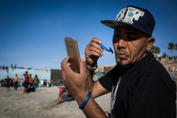 Ein Flüchtling aus Honduras rasiert sich am 13. November 2018 in Tijuana (Mexiko) im Freien. Im Hintergrund stehen Menschen an dem Grenzzaun zu den USA.