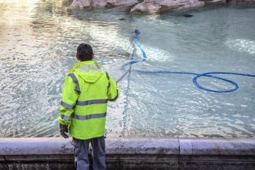Ein Mann schiebt am 15. März 2019 Münzen in die Mitte des Trevibrunnens in Rom. Ein umgebauter Poolsauger saugt Münzen vom Grund.
