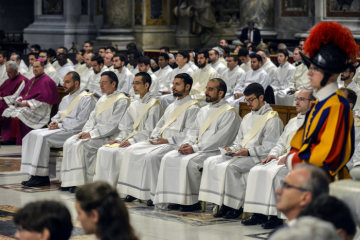 Die Weihekandidaten in weißen Alben sitzen auf einer Kirchenbank am 12. Mai 2019 im Petersdom im Vatikan. Neben ihnen steht ein Schweizergardist.