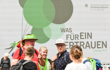Menschen stehen bei der Eröffnung des Deutschen Evangelischen Kirchentags (DEKT) am 19. Juni 2019 in Dortmund vor einer Plakatwand mit dem Motto und dem Logo des Kirchentags.