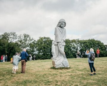 Statue des Heiligen Moe (Saint Moe) aus Granit im Tal der Heiligen am 25. August 2020 in Carnoet. Dahinter weitere Granitstatuen, dazwischen laufen Touristen umher.