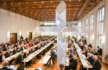 Blick durch ein gelochtes Metallkreuz auf die Teilnehmer an langen Tischen bei den Beratungen der Synodalversammlung am 31. Januar 2020 im Dominikanerkloster in Frankfurt.