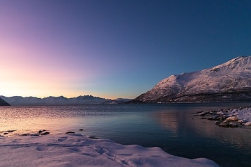 Das wenige Tageslicht über einer schneebedeckten Bergkette schimmert rötlich-perlmuttfarben bis blau und spiegelt sich im Wasser einer Meeresbucht am 14. Dezember 2023 in der Nähe von Alta (Norwegen).