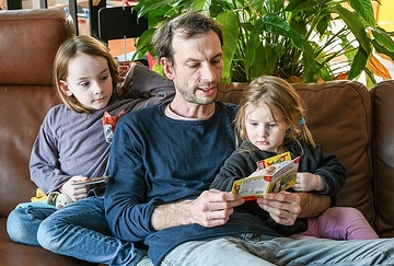 Vater, Tochter und Sohn (l.) sitzen am 6. März 2024 auf dem Sofa zuhause in Rheinbach. Der Vater liest den Kindern aus einem Pixi-Buch vor.