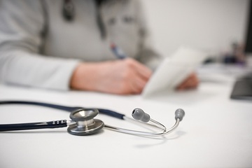 Ein Stethoskop liegt auf einem Schreibtisch im Sprechzimmer einer Arztpraxis am 21. Dezember 2022 in Bonn. Eine Ärztin macht Notizen in einer Krankenakte.