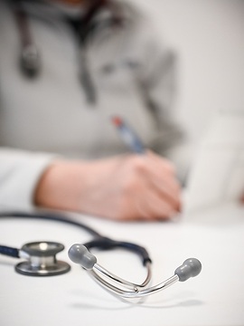 Ein Stethoskop liegt auf einem Schreibtisch im Sprechzimmer einer Arztpraxis am 21. Dezember 2022 in Bonn. Eine Ärztin macht Notizen in einer Krankenakte.
