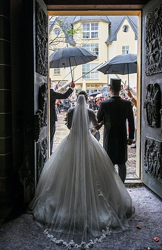 Ein Brautpaar verlässt nach der Trauung die Kirche durch das Hauptportal, am 20. April 2024 in der Kirche Sankt Laurentius in Bad Neuenahr-Ahrweiler. Auf dem Boden liegt Malervlies. Zwei Gäste schützen das Paar mit Regenschirmen vor dem Regen.