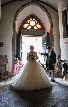 Die Braut steht vor Beginn des Gottesdienstes im Hauptportal auf einem Malervlies, am 20. April 2024 in der Kirche Sankt Laurentius in Bad Neuenahr-Ahrweiler. Zwei Gäste richten das weiße Brautkleid.